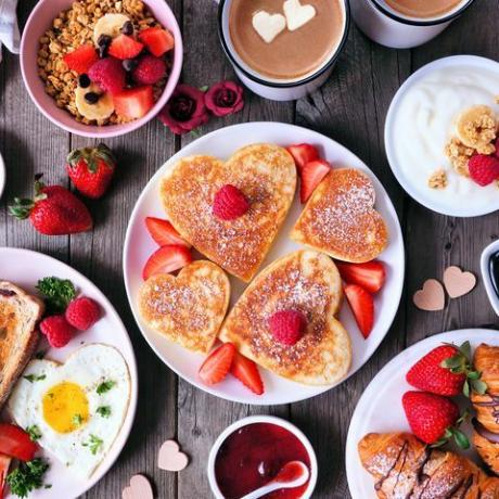 Valentinstag oder Muttertagsfrühstückstischszene auf dunklem Holzhintergrund mit herzförmigen Pfannkuchen, Eiern und Liebesspeisen