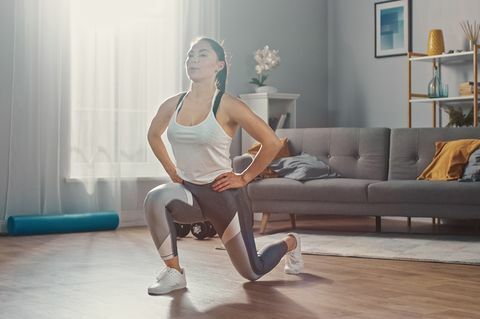 Starkes und schönes athletisches Fitness-Mädchen in Sportbekleidung macht Vorwärts-Longe-Übungen in ihrem hellen und geräumigen Wohnzimmer mit minimalistischem Interieur.